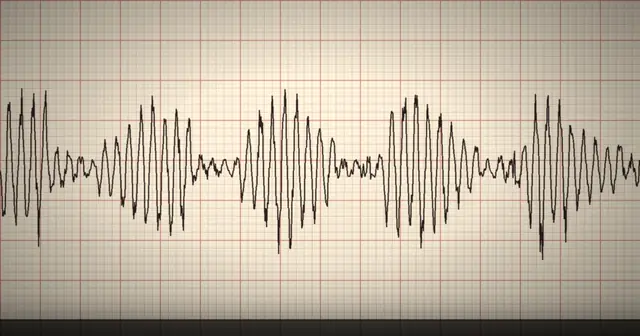 A strip of heated chart paper with a jagged black voice-stress trace drawn across a red grid